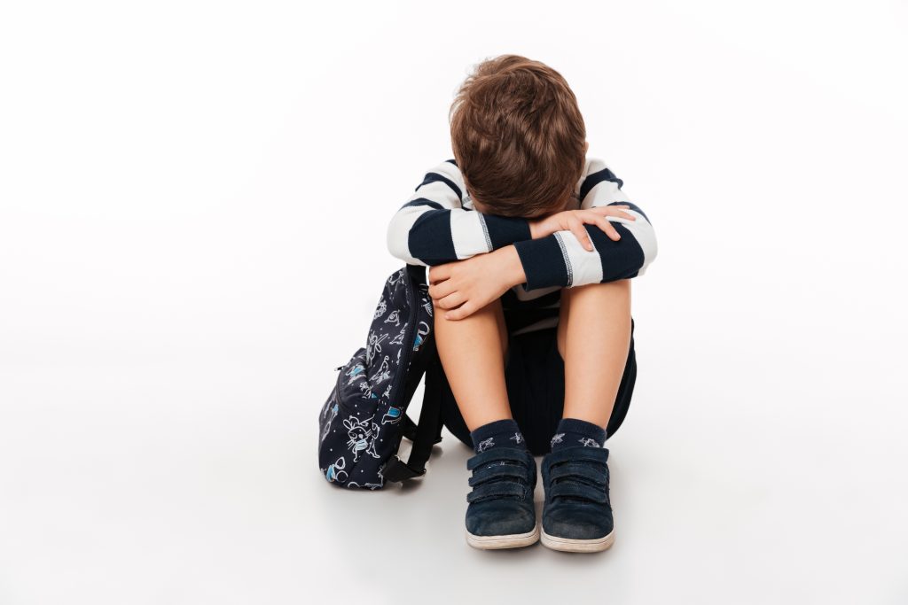 Portrait of an upset sad little kid with backpack sitting on a flor and crying isolated over white background